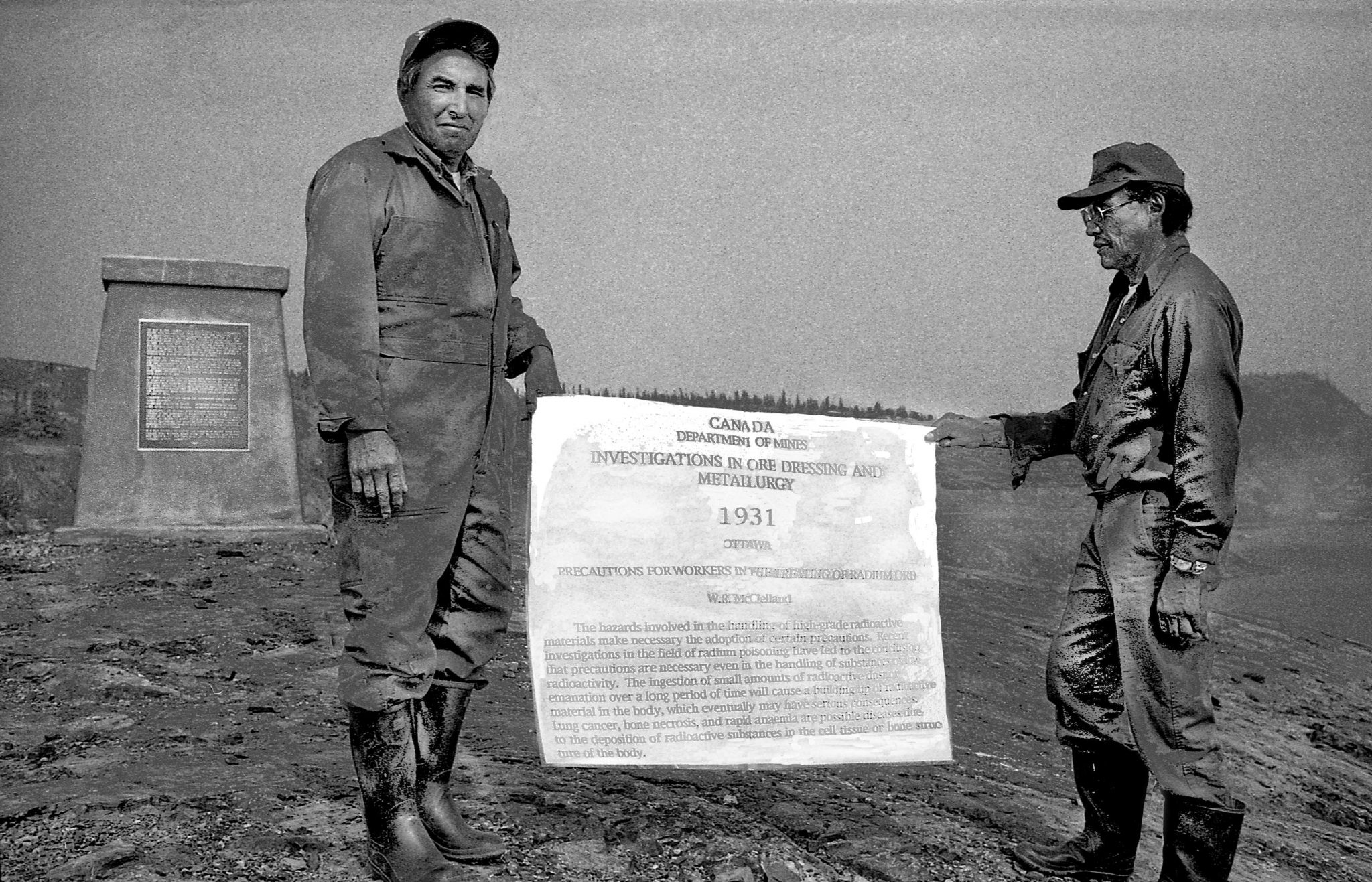 Two former Dene uranium ore carriers at the Port Radium mine on Great Bear Lake hold up an advisory published by the Canadian Department of Mines in 1931 describing the hazards of handling uranium ores from Port Radium or any other uranium mine. The bronze plaque on the cairn in the background describes the kind of work done at Port Radium.